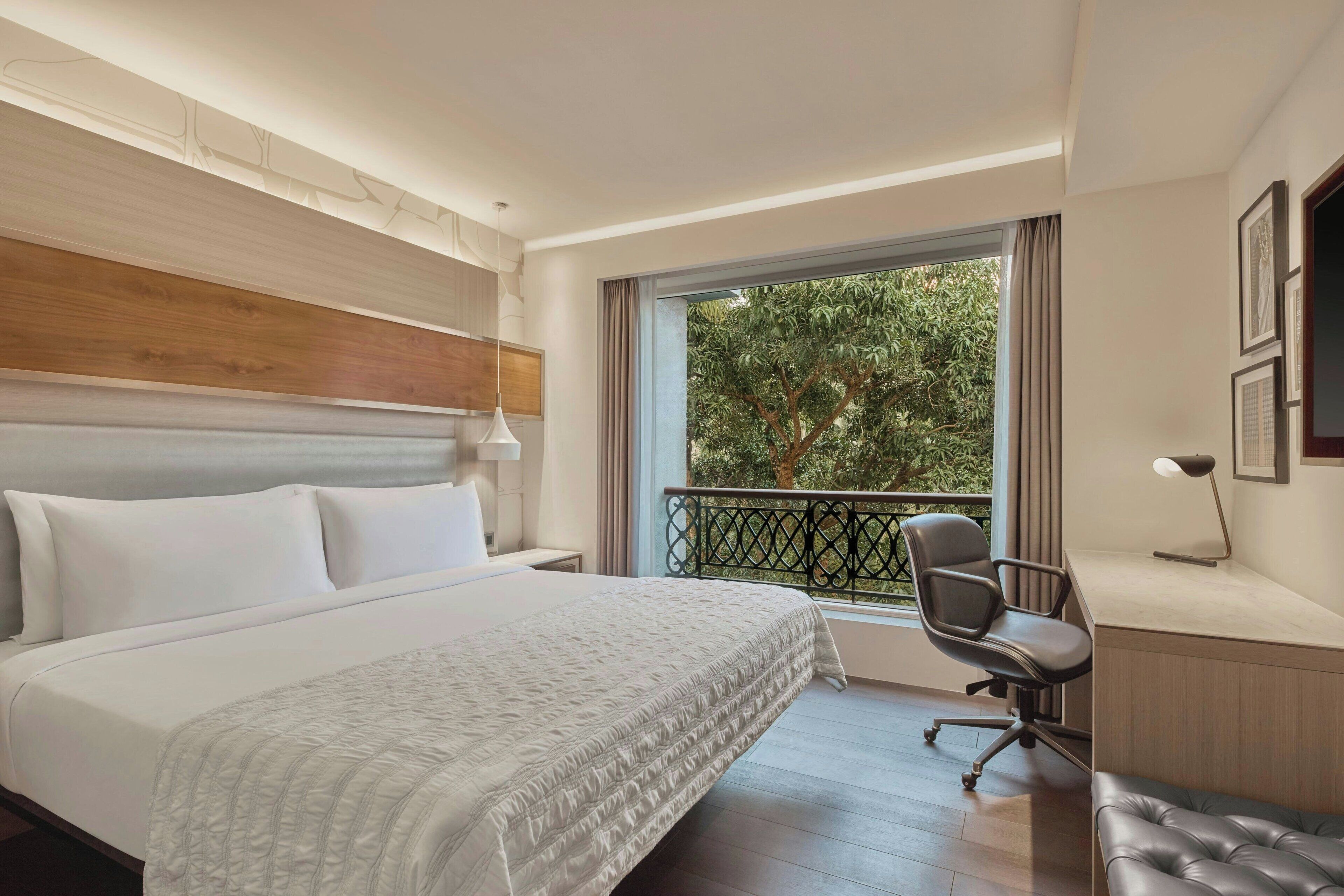 A hotel room with a large bed, a desk with a chair, a balcony with a view of trees, and a TV mounted on the wall.