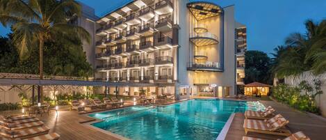 A hotel with a swimming pool, lounge chairs, and a well-lit building facade.