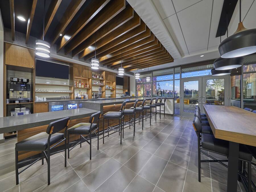 A modern bar area with multiple seating options, including bar stools and high tables, and a kitchen area with visible appliances and shelves stocked with bottles.