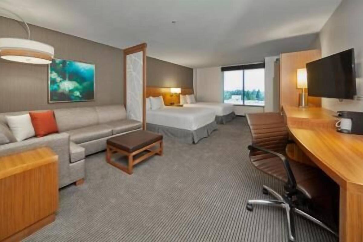 A hotel room with a sofa, a bed, a desk with a chair, a TV, and a painting on the wall.
