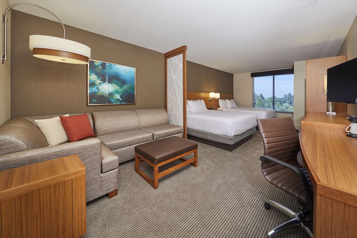 A hotel room with a sofa, two single beds, a desk with a coffee maker, a TV, and a large window with a view.