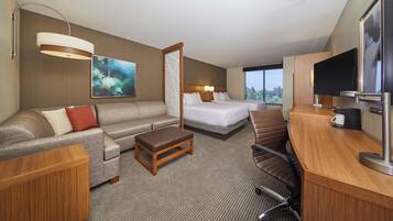 A hotel room with a sofa, two single beds, a desk with a coffee maker, a TV, and a large window with a view.