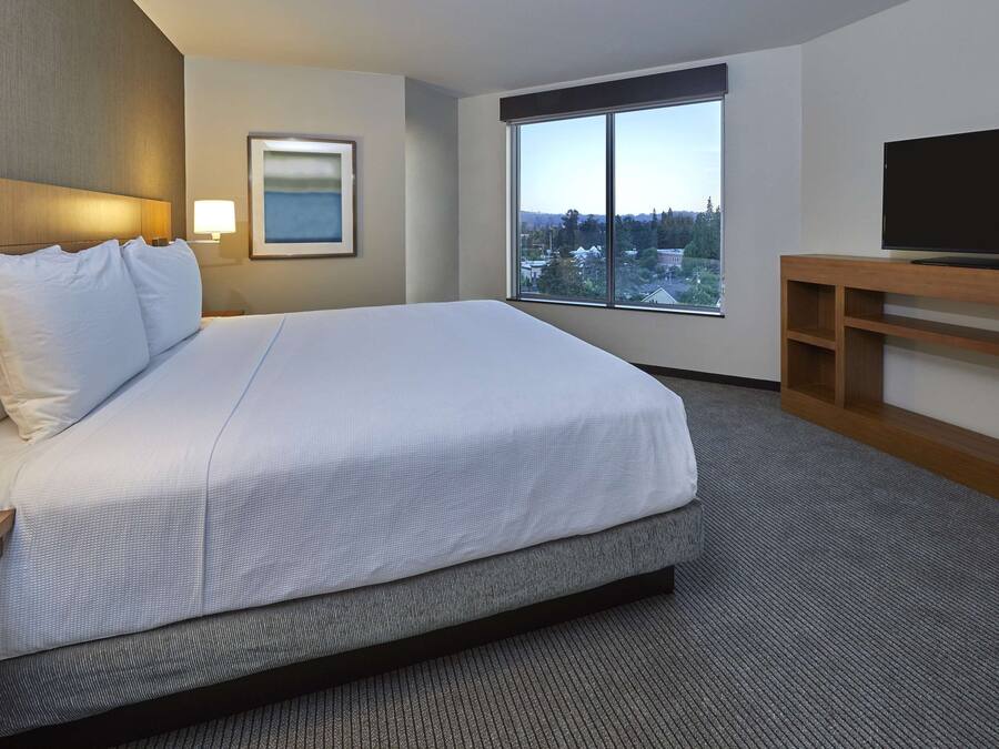 Ein Hotelzimmer mit einem großen Bett, einem Fernseher auf einem Holztisch, einem Fenster mit Blick auf die Stadt und einer Lampe auf dem Nachttisch.