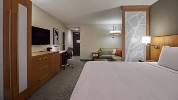 A hotel room with a large bed, a desk with a chair, a TV, a sofa, and a nightstand with a lamp.