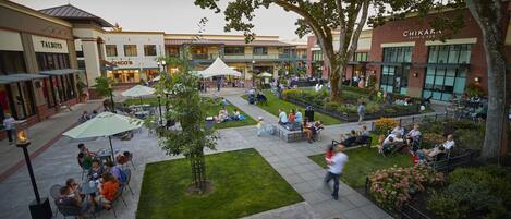 An outdoor plaza with seating areas, trees, and a variety of shops in the background.