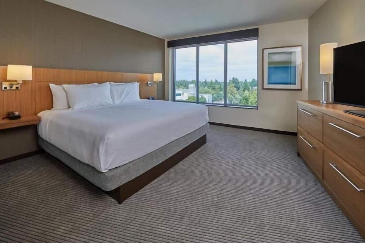 A hotel room with a bed, a TV, a desk with drawers, a window with a view, and a wall-mounted lamp.