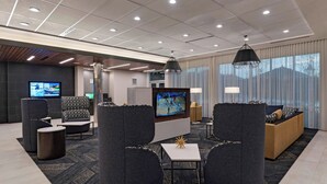 A modern hotel lobby with grey upholstered seating, a flat-screen TV, and a large window with curtains. - Courtyard by Marriott Houston Sugar Land/Lake Pointe (Sugar Land)