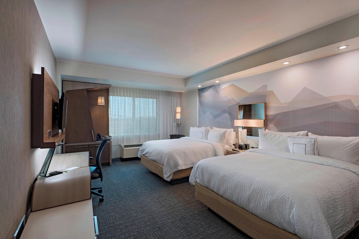 A hotel room with two beds, a desk, a chair, a TV, and a wall-mounted mirror.