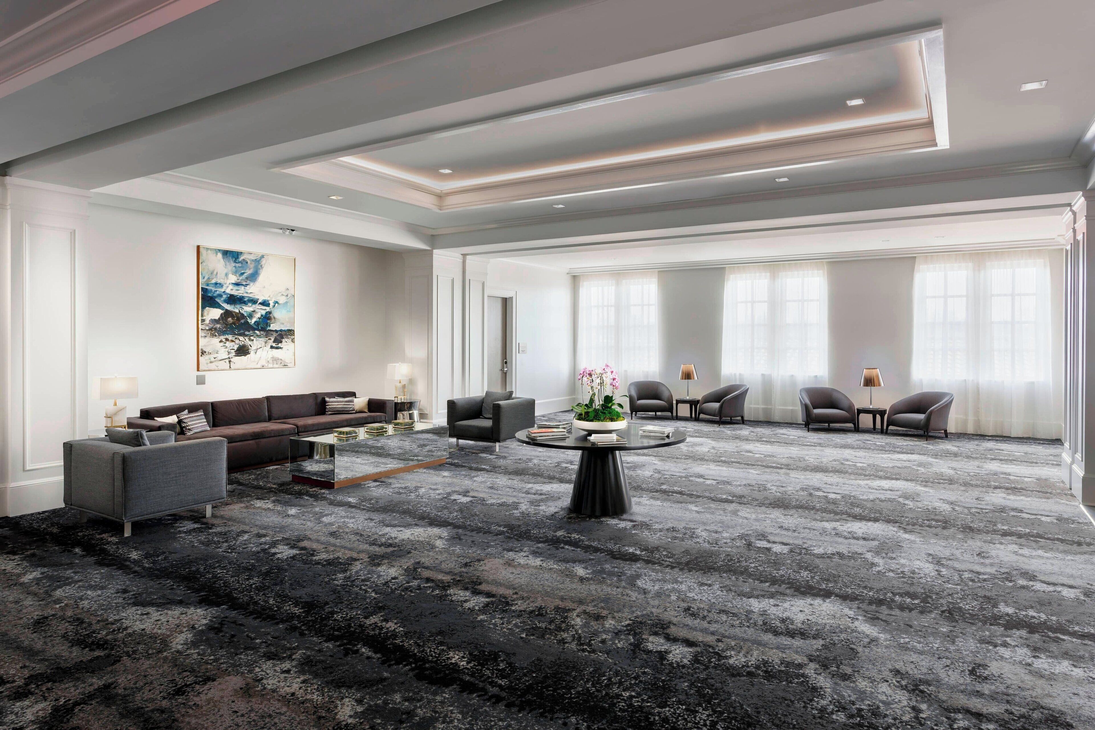 A spacious living room with a large black and gray carpet, a brown sofa, a round coffee table with a plant, and a large abstract painting on the wall.