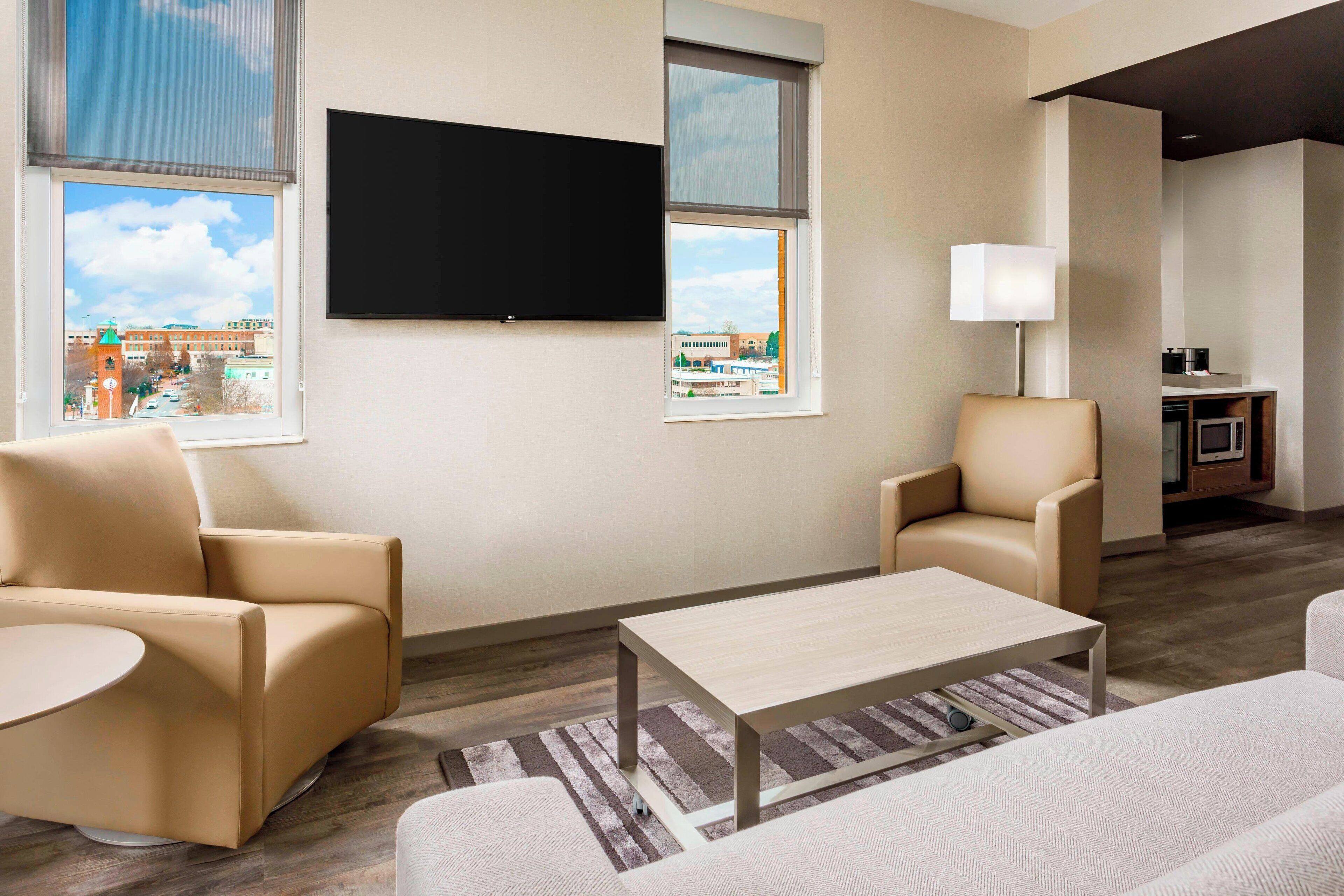 Una sala de estar moderna con un televisor de pantalla plana, dos sofás, una mesa de centro y vistas a edificios a través de ventanas amplias.