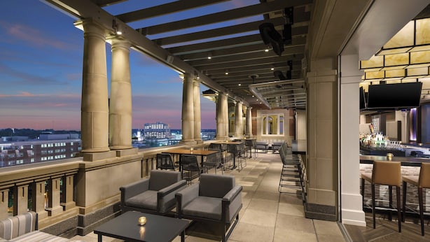 Een terras op het dak met zitplaatsen, een bar en een uitzicht op de stad in de schemering.