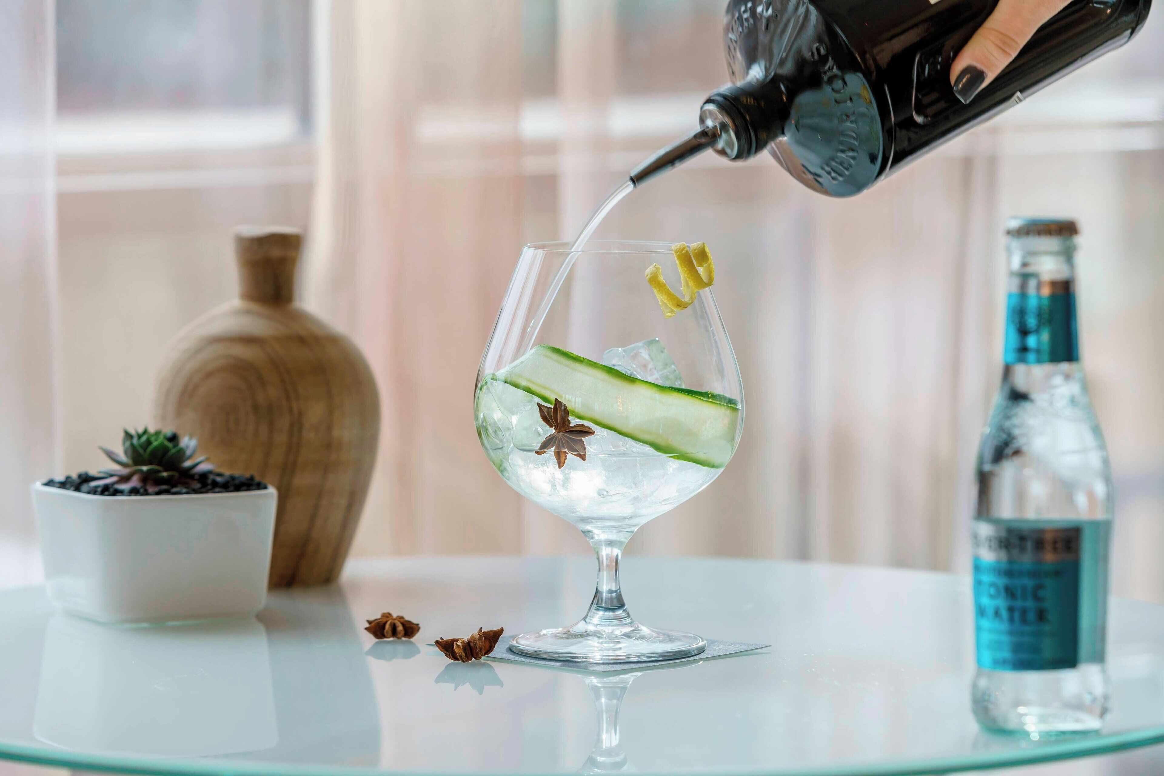 A glass with ice, cucumber slices, and star anise being filled with water from a bottle.