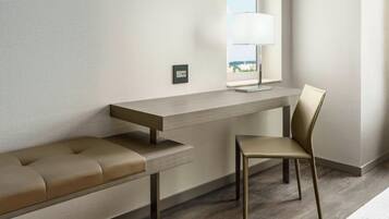 A modern hotel room with a wooden desk, a cushioned bench, a chair, and a window with a view.