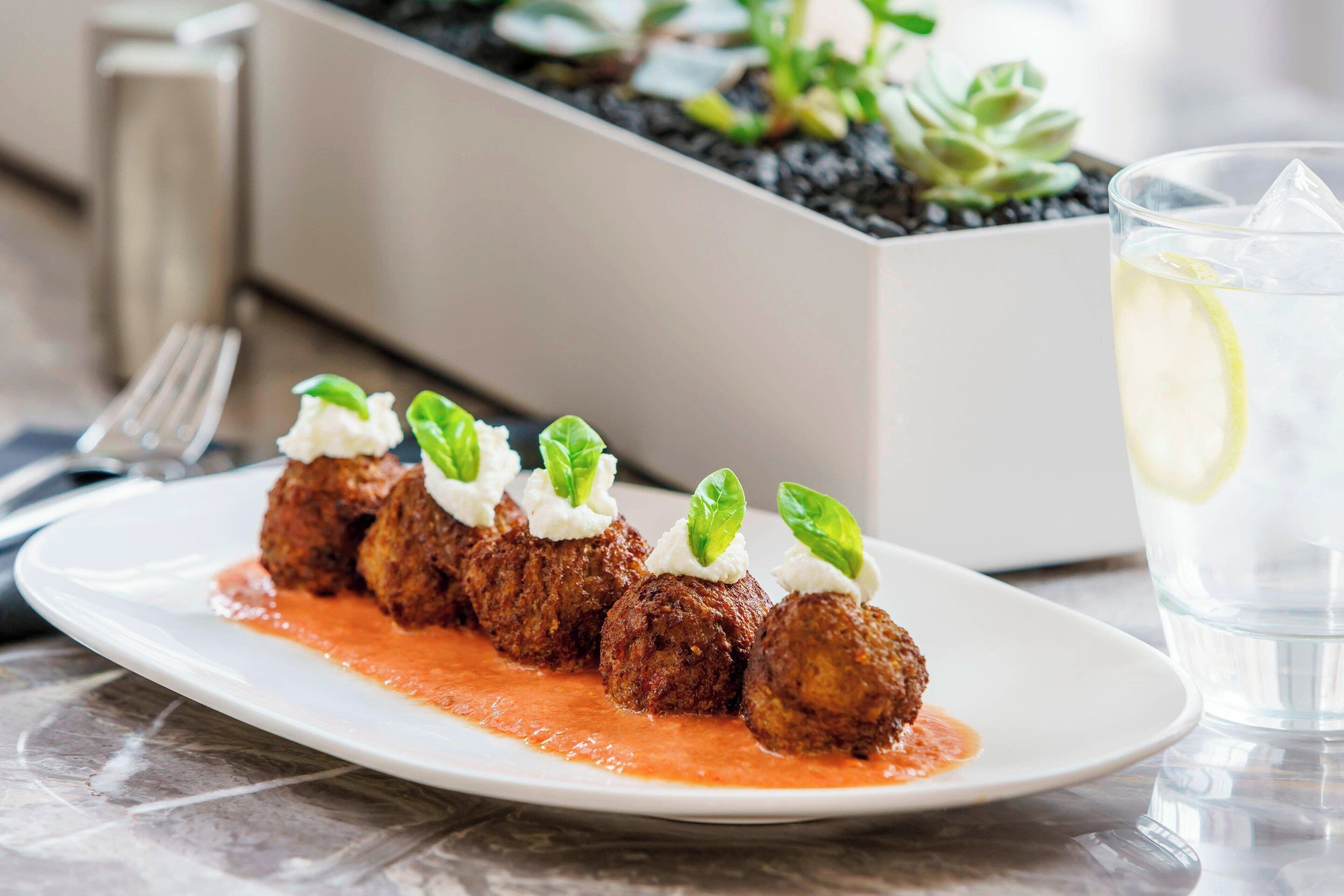 Un plato de albóndigas con salsa y decoración, un vaso de agua con rodaja de limón y una planta al fondo.