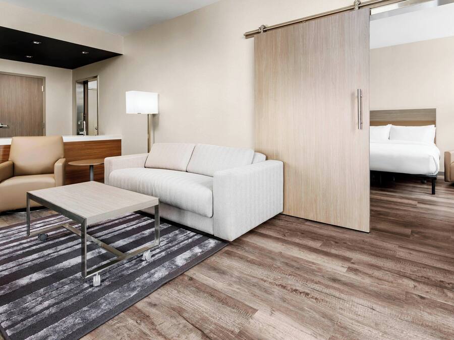 Una habitación de hotel moderna con sofá, una mesita, una lámpara y una puerta corredera que da a un dormitorio.