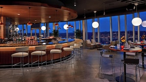 A modern bar area with a curved counter, multiple bar stools, and large windows offering a city view at dusk. - AC Hotel by Marriott Columbus Dublin (Dublin)