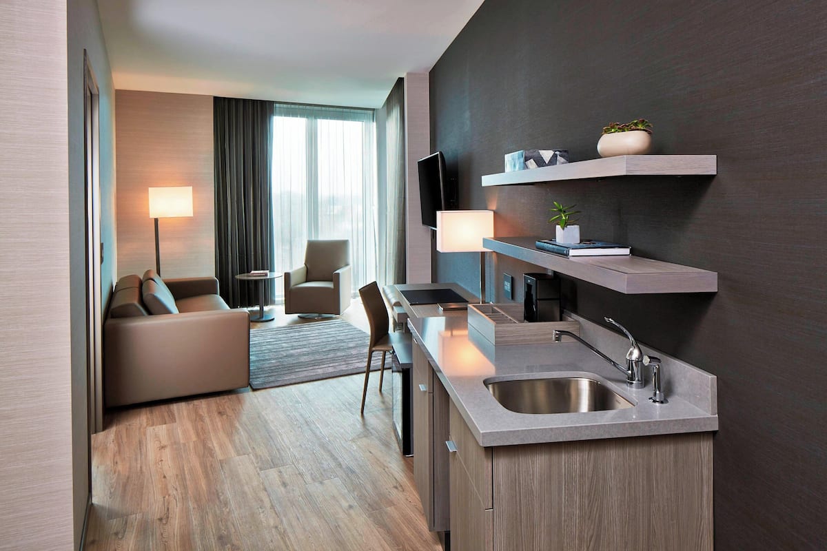 Una habitación de hotel moderna con kitchenette, un sofá, zona de comedor y televisor montado en la pared.