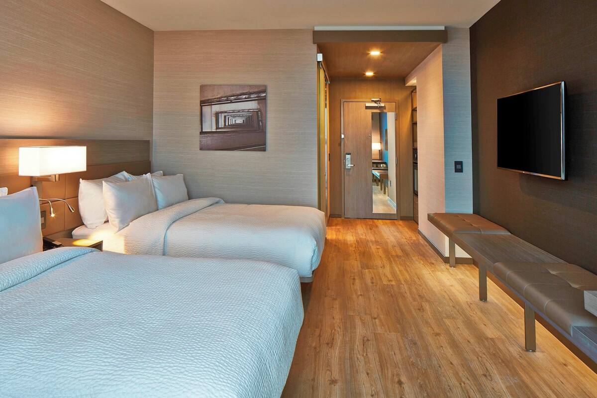 Habitación de hotel con dos camas, un banco, un televisor y un pasillo que conduce a un baño.