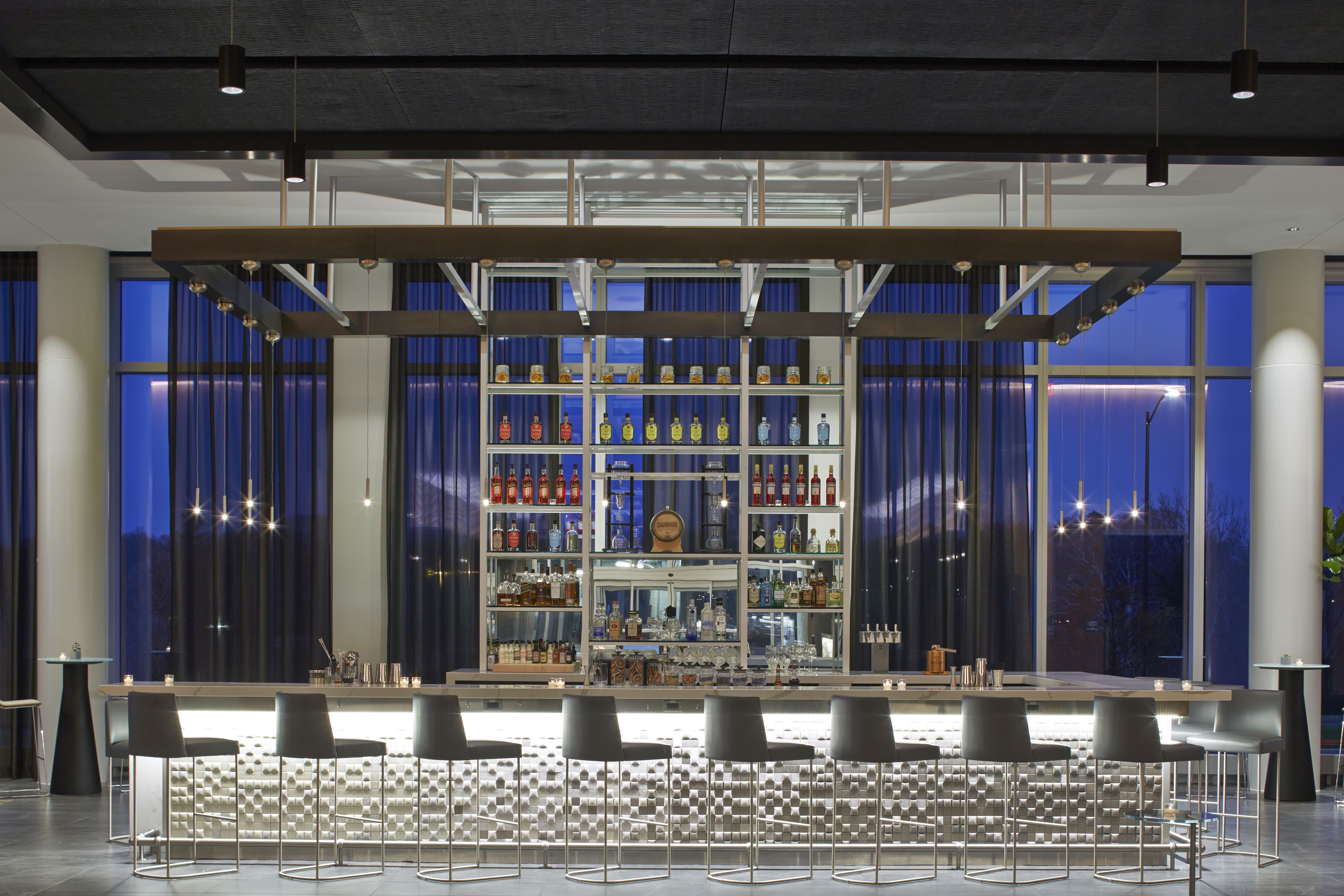 A bar area with a central shelf displaying various bottles, bar stools, and a large window with curtains.
