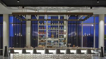 A bar area with a central shelf displaying various bottles, bar stools, and a large window with curtains.