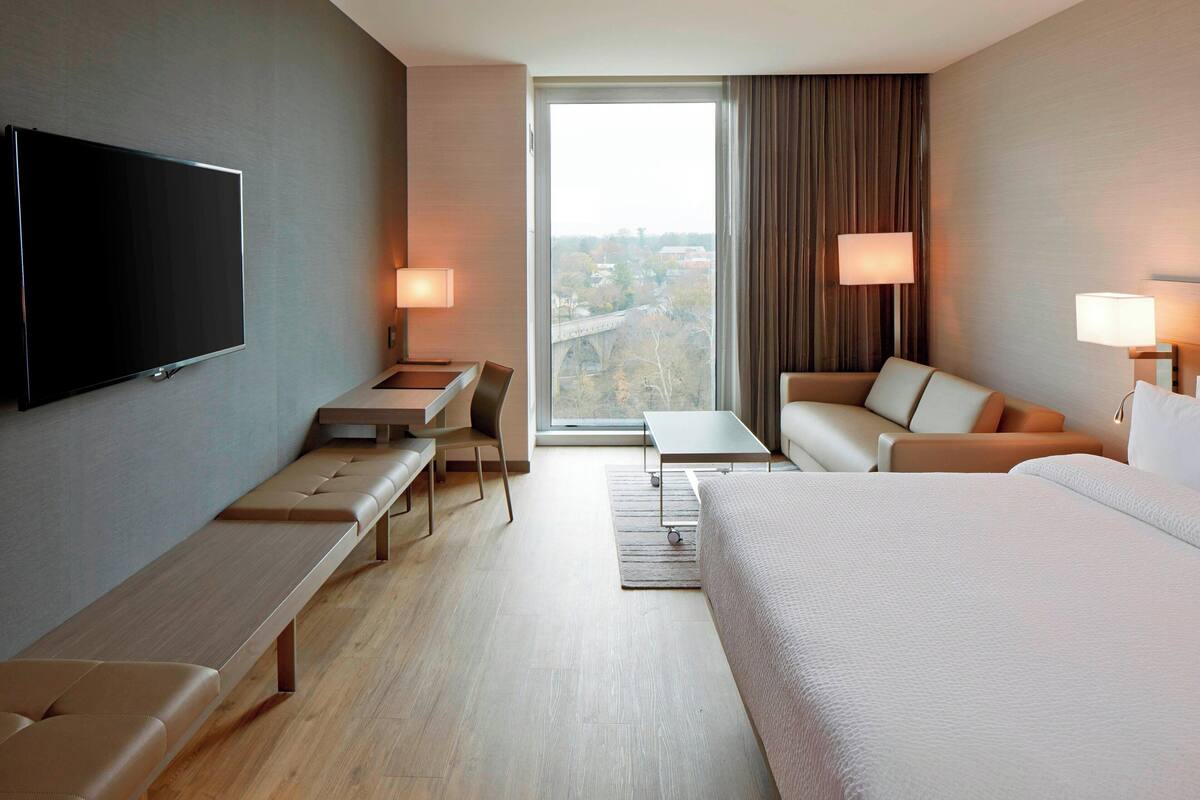 Una habitación de hotel moderna con una cama grande, un escritorio con una silla, un banco, un televisor montado en la pared y un amplio ventanal con vistas a la ciudad.