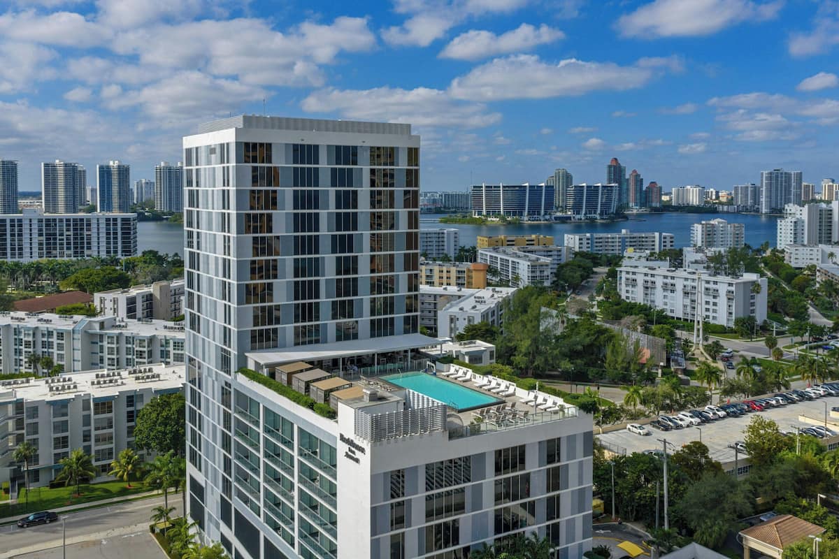 A modern high-rise hotel with a rooftop pool, surrounded by a cityscape and a body of water.
