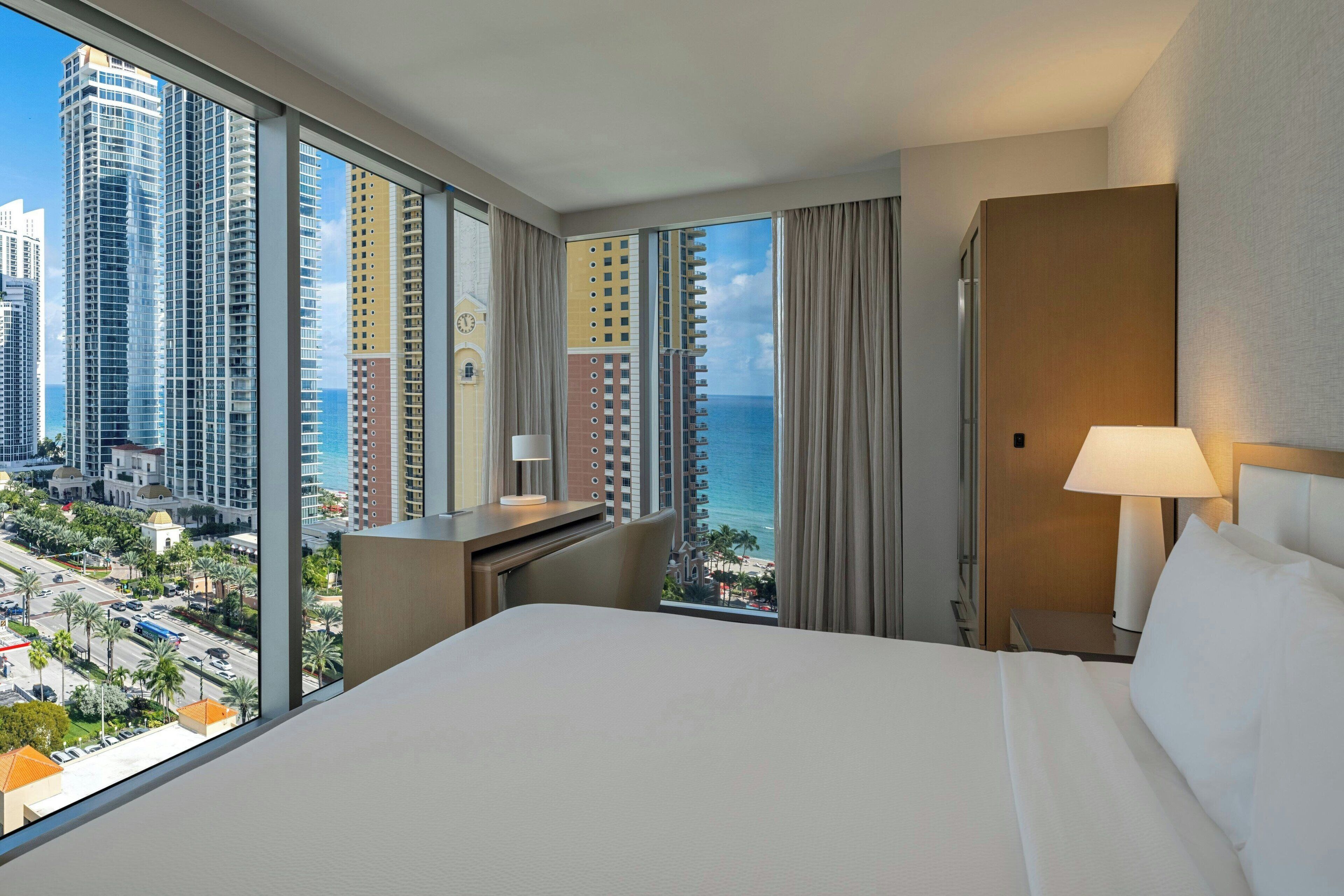 Ein Hotelzimmer mit einem großen Bett, einem Schreibtisch und Blick auf Stadt und Strand.