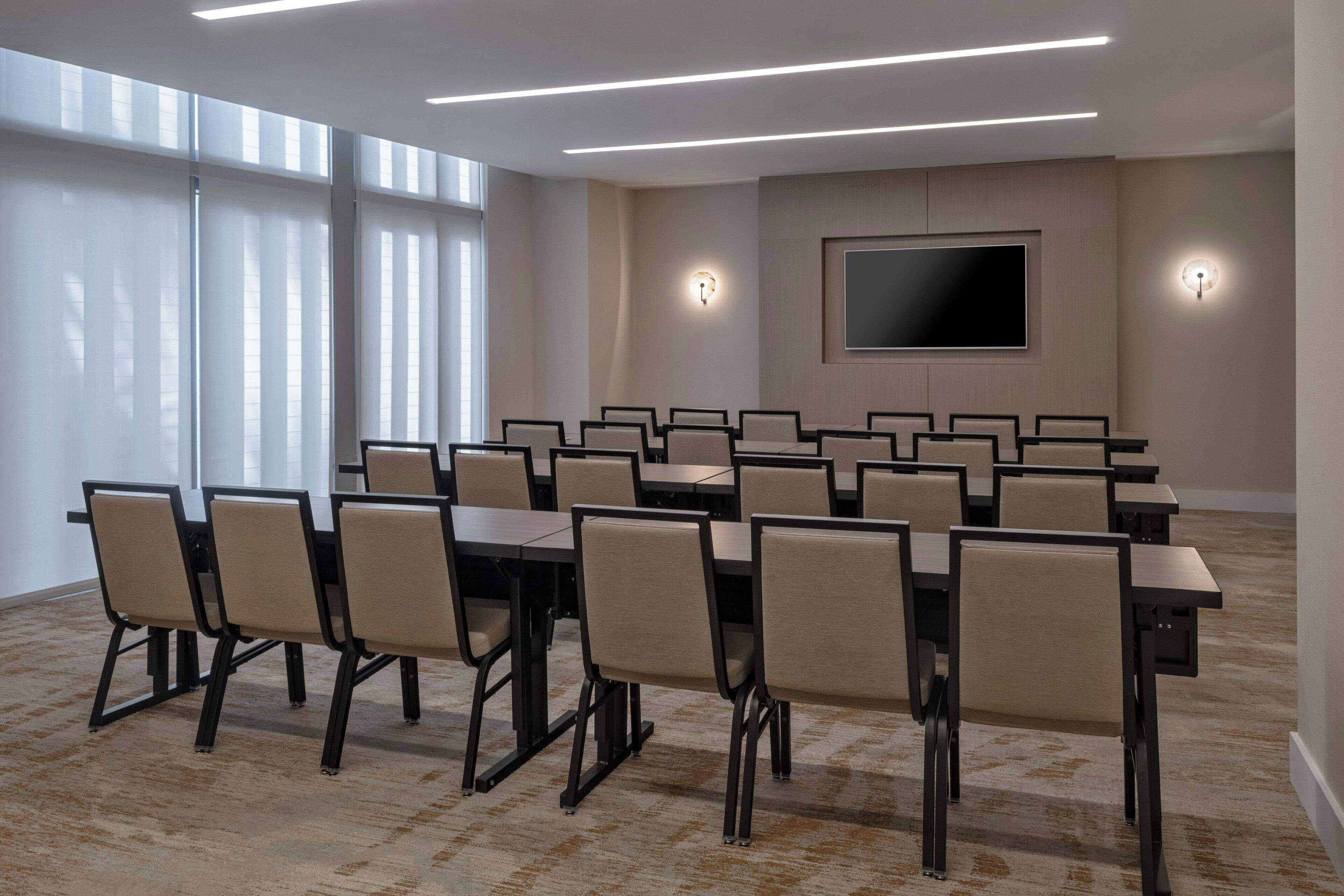 Une salle de conférence équipée de plusieurs tables et chaises, d’un grand téléviseur à écran plat fixé au mur et de deux luminaires muraux.