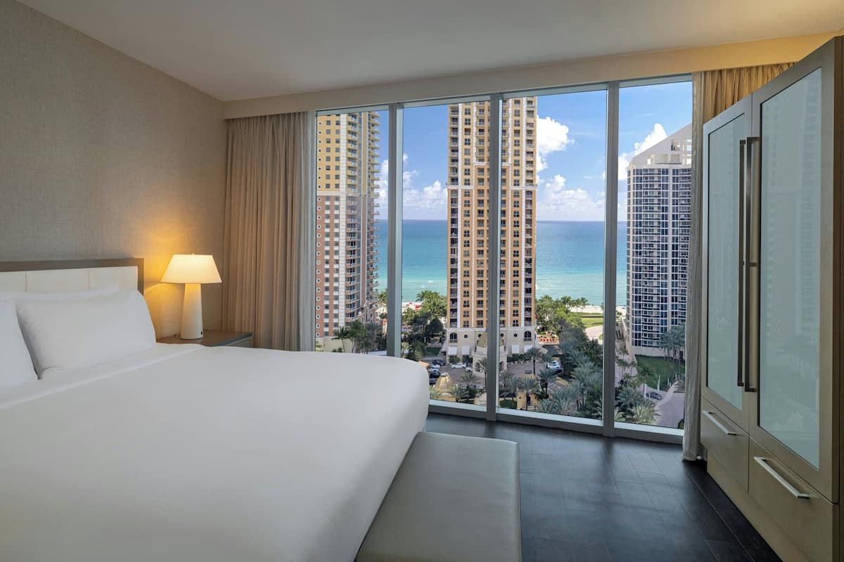 A hotel room with a large bed, a nightstand with a lamp, a wardrobe, and a view of tall buildings and the ocean.