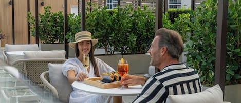 A couple seated at an outdoor table with a glass of drink and a tray of food, surrounded by greenery and modern outdoor furniture.