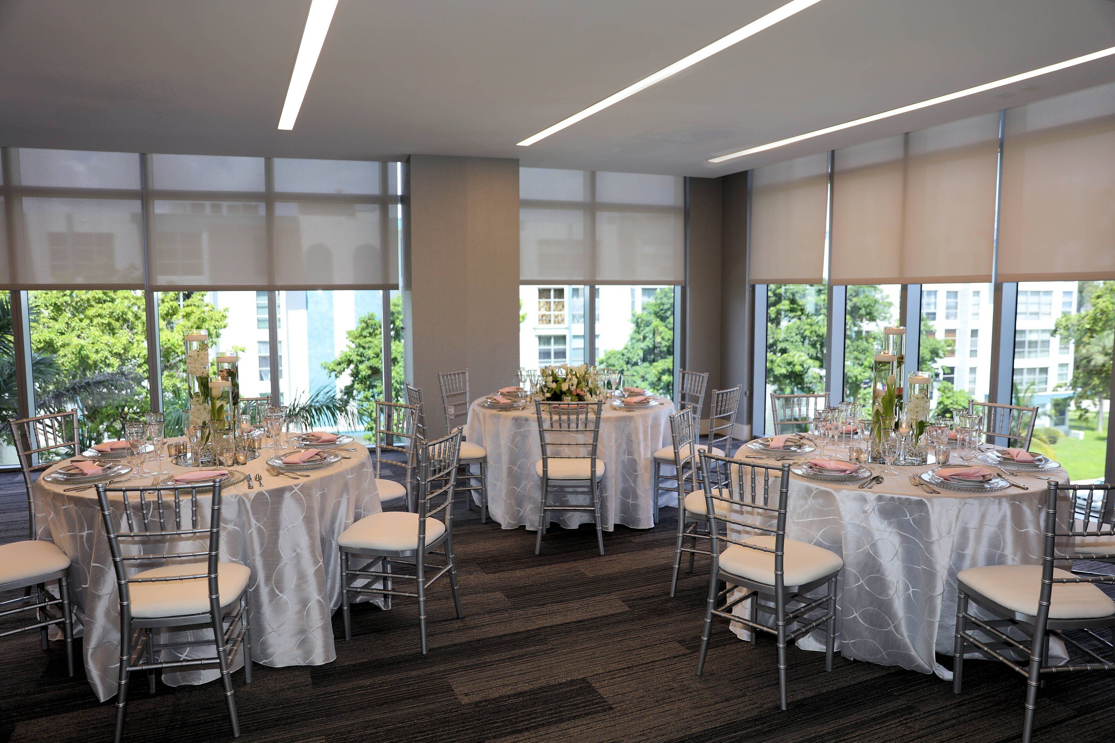 Een feestzaal met ronde tafels gedekt voor een formeel evenement, grote ramen met uitzicht op de stad, en een vloerbedekt met tapijt.