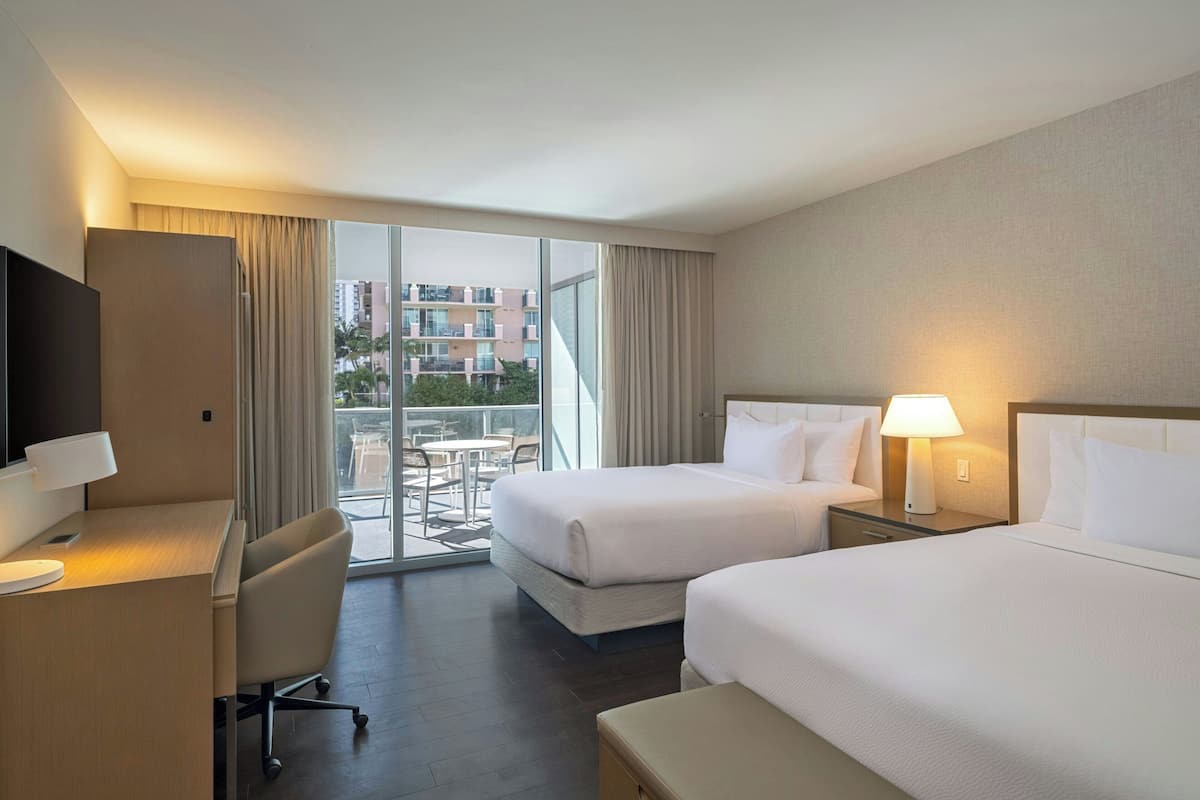 A hotel room with two beds, a desk, a chair, a TV, and a balcony with a view of buildings.