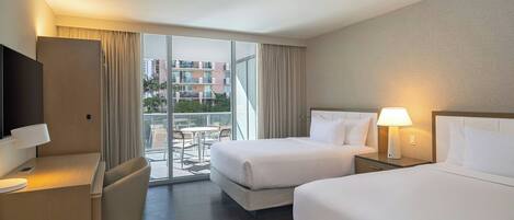 Habitación de hotel con dos camas, un escritorio, una silla, un televisor y un balcón con vista a edificios.