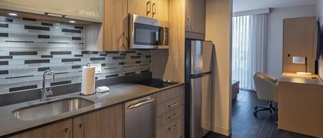 A modern kitchen with a stainless steel microwave, refrigerator, and sink. There is a wooden cabinet and a tiled backsplash. A chair and a desk are visible in the adjacent room.