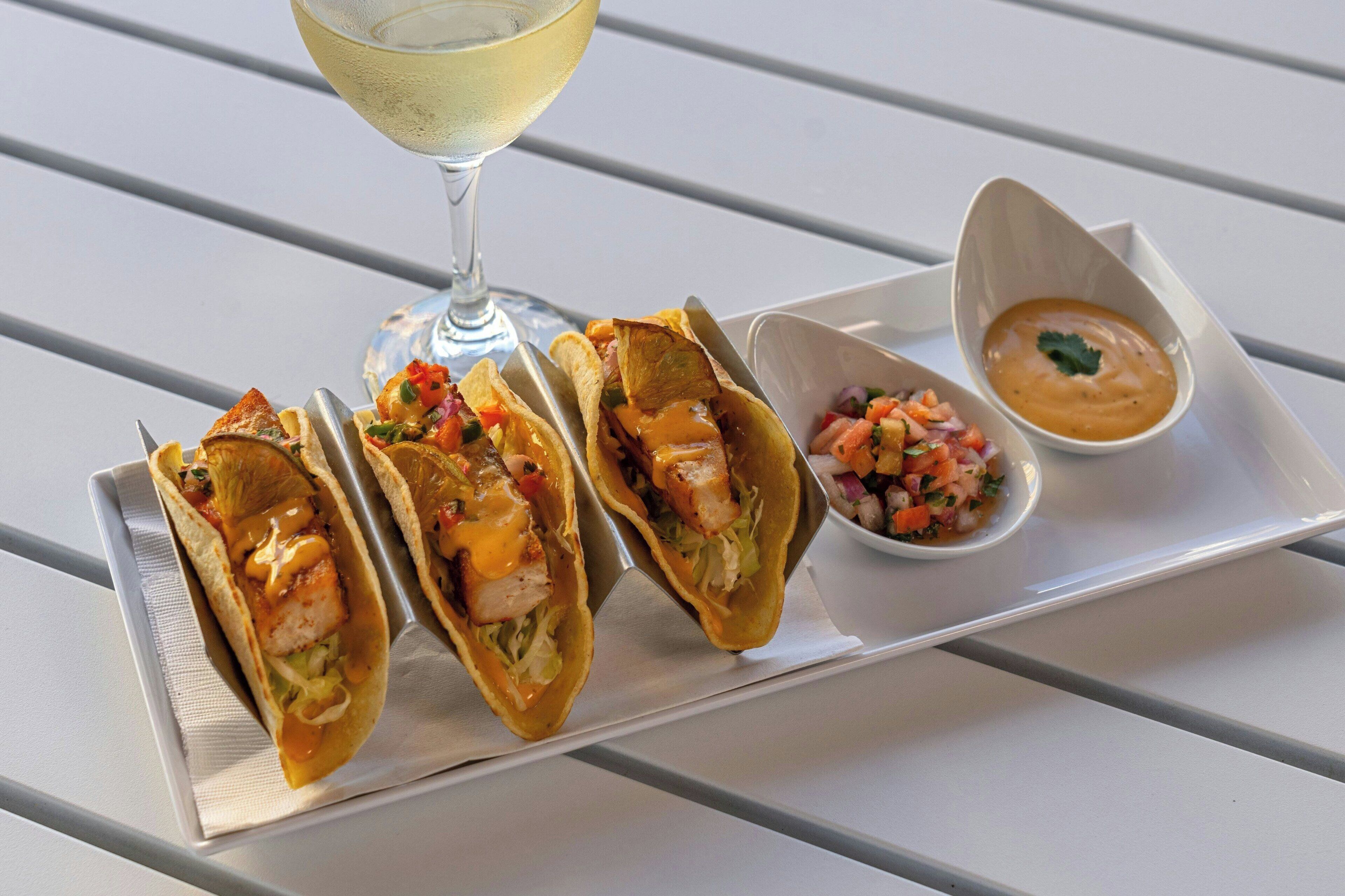 Three tacos on a white tray with a glass of white wine and two small bowls of salsa and sauce.