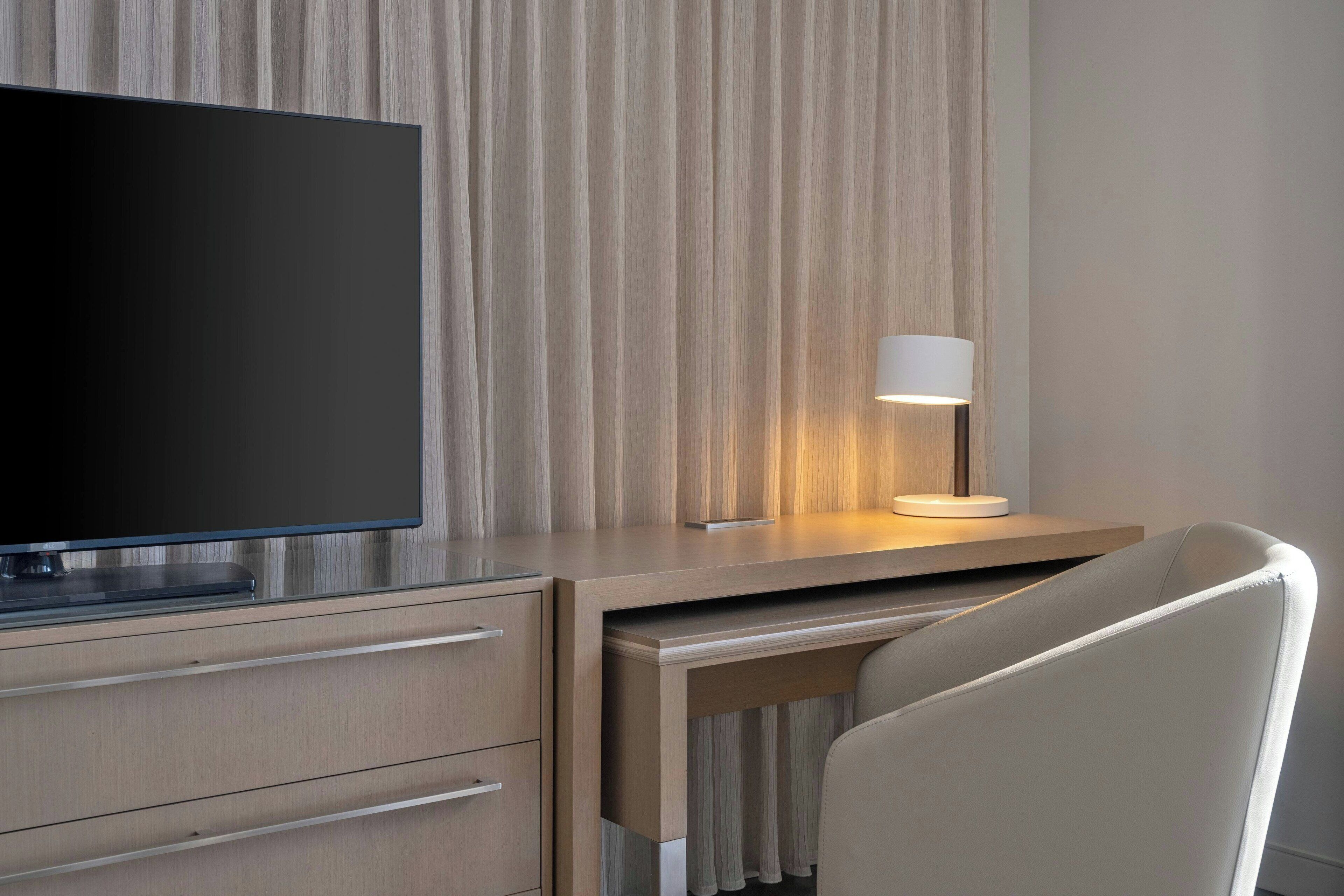 A modern hotel room with a flat-screen TV, a desk with a lamp, and a chair.