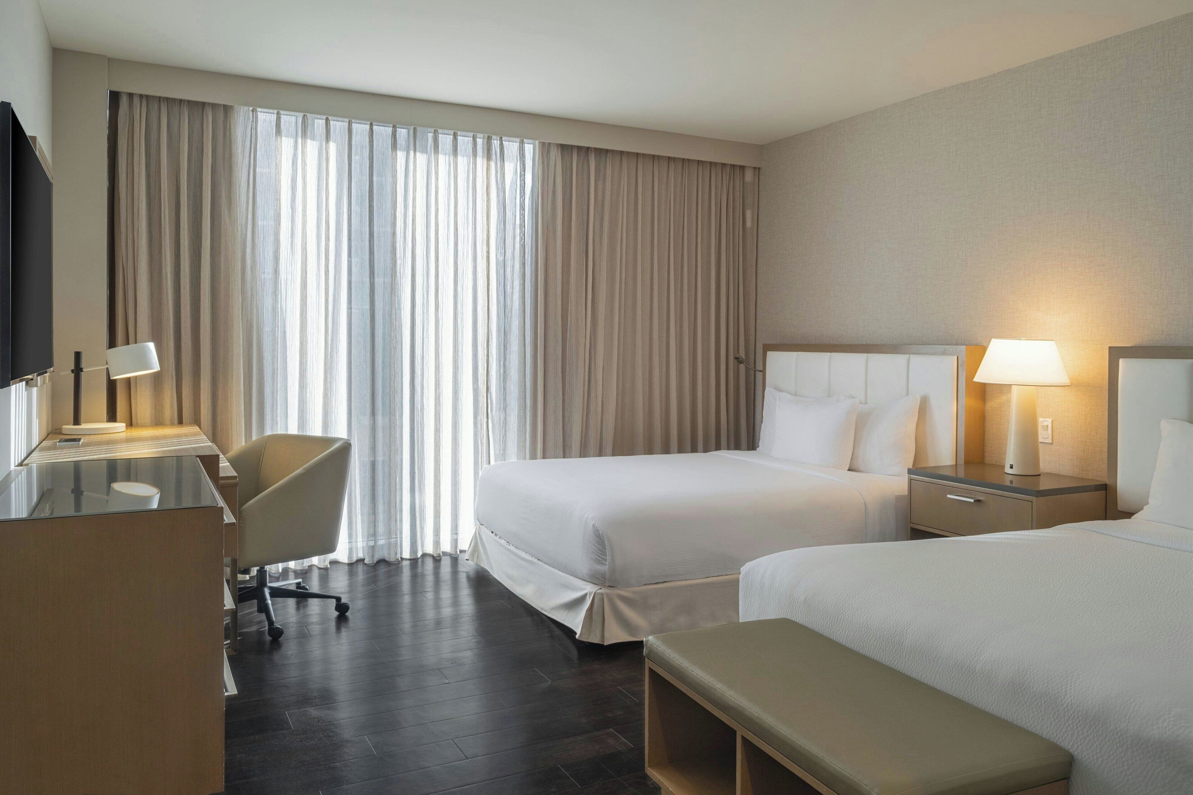 A hotel room with two beds, a desk, a chair, and a TV.