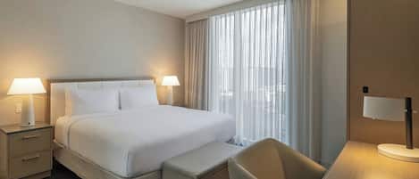 A hotel room with a large bed, two bedside lamps, a desk with a chair, and a view of the city through sheer curtains.