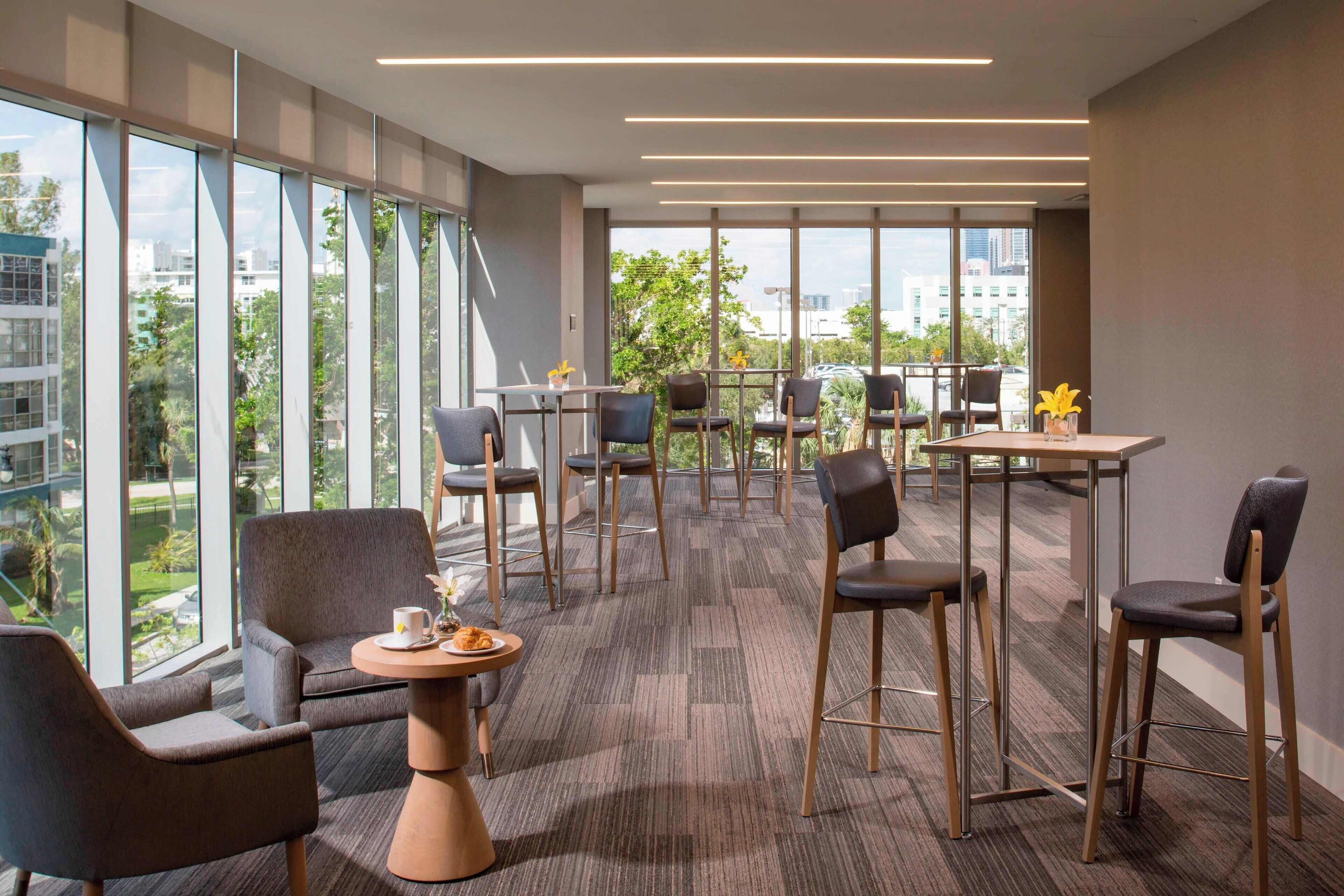 A modern hotel lobby with a high ceiling, large windows offering a city view, and a mix of seating arrangements including chairs and tables.