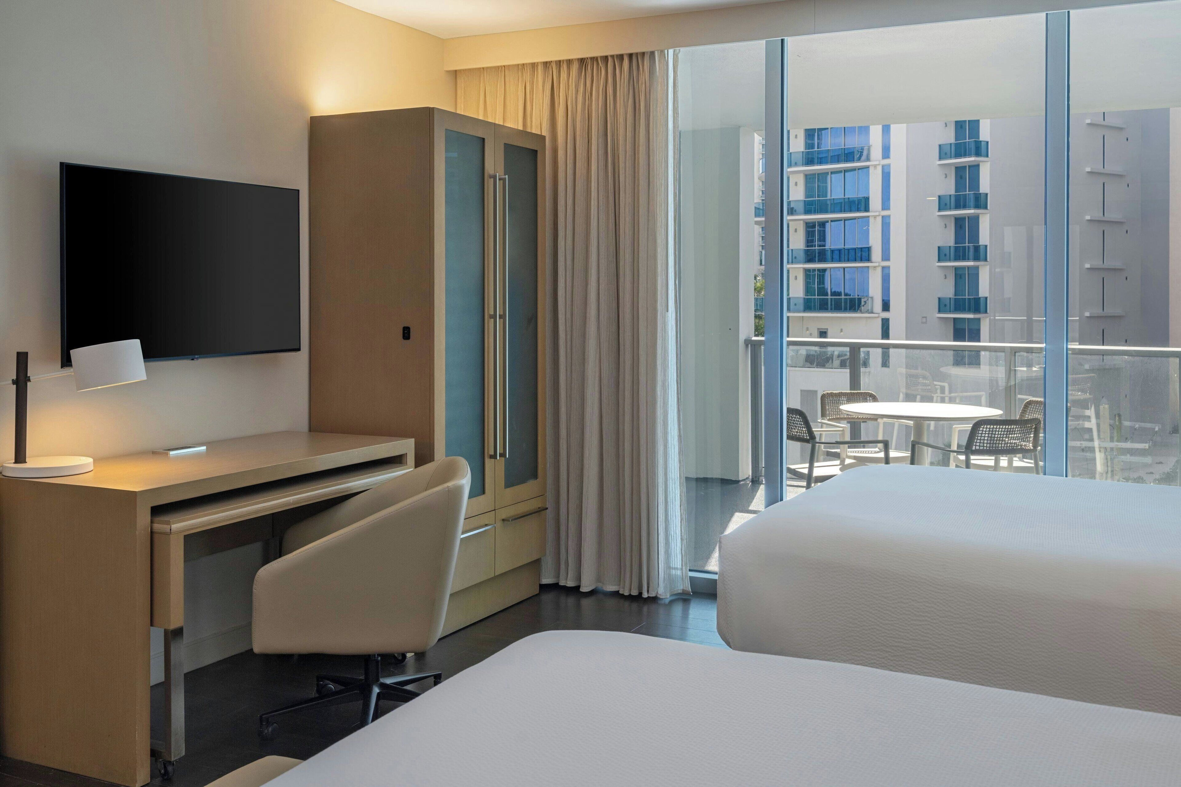 A hotel room with a bed, a desk, a chair, a TV, and a balcony with a view of buildings.