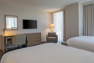 Quarto de hotel com cama, escrivaninha com cadeira, televisão e luminária.