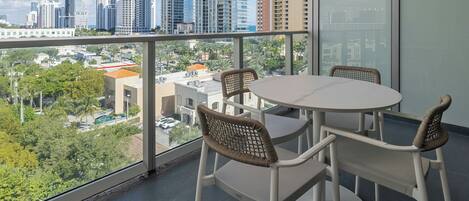 A balcony with a round table and wicker chairs, overlooking a cityscape with tall buildings and palm trees.