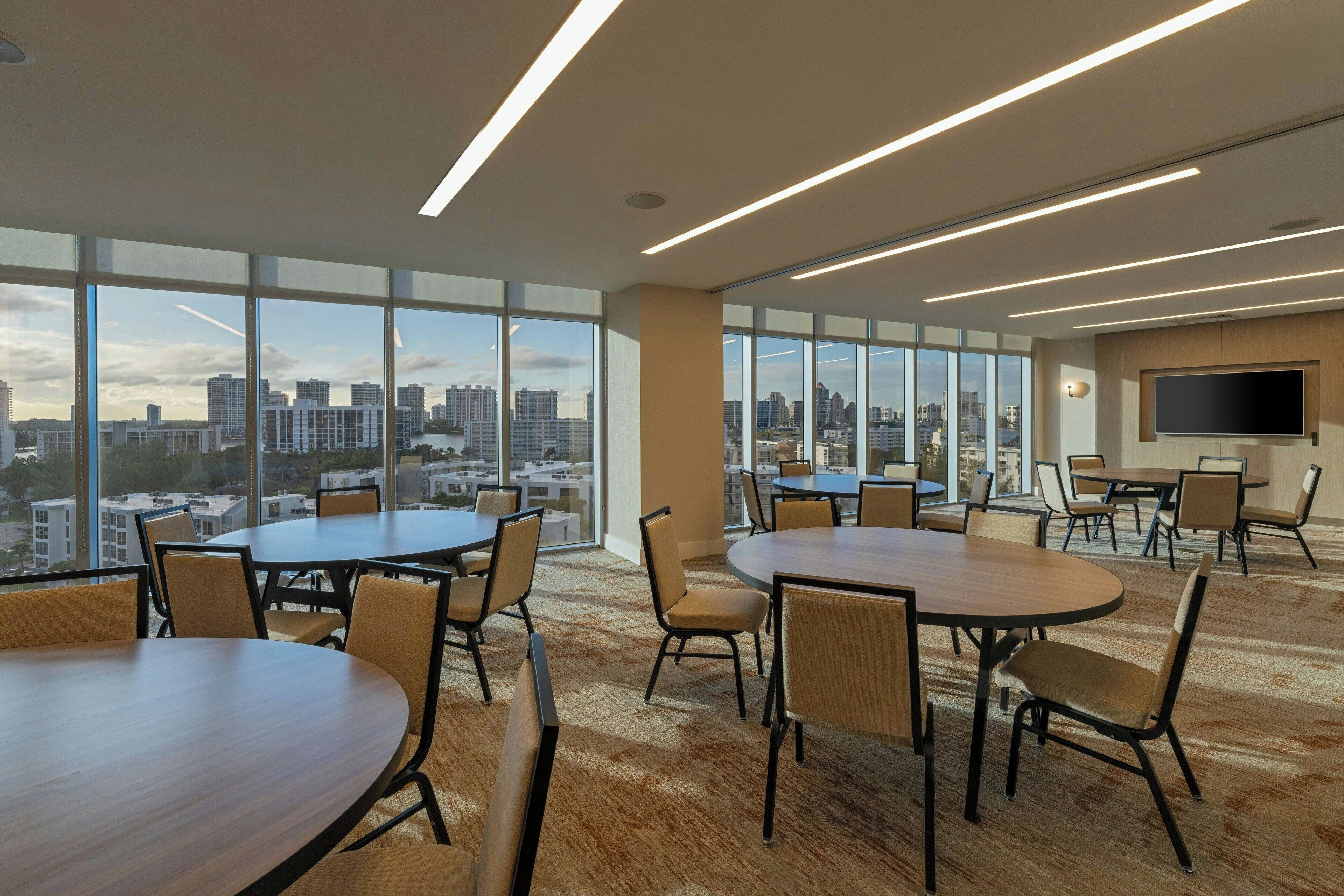 A modern conference room with large windows, multiple tables, and chairs, and a flat-screen TV mounted on the wall.