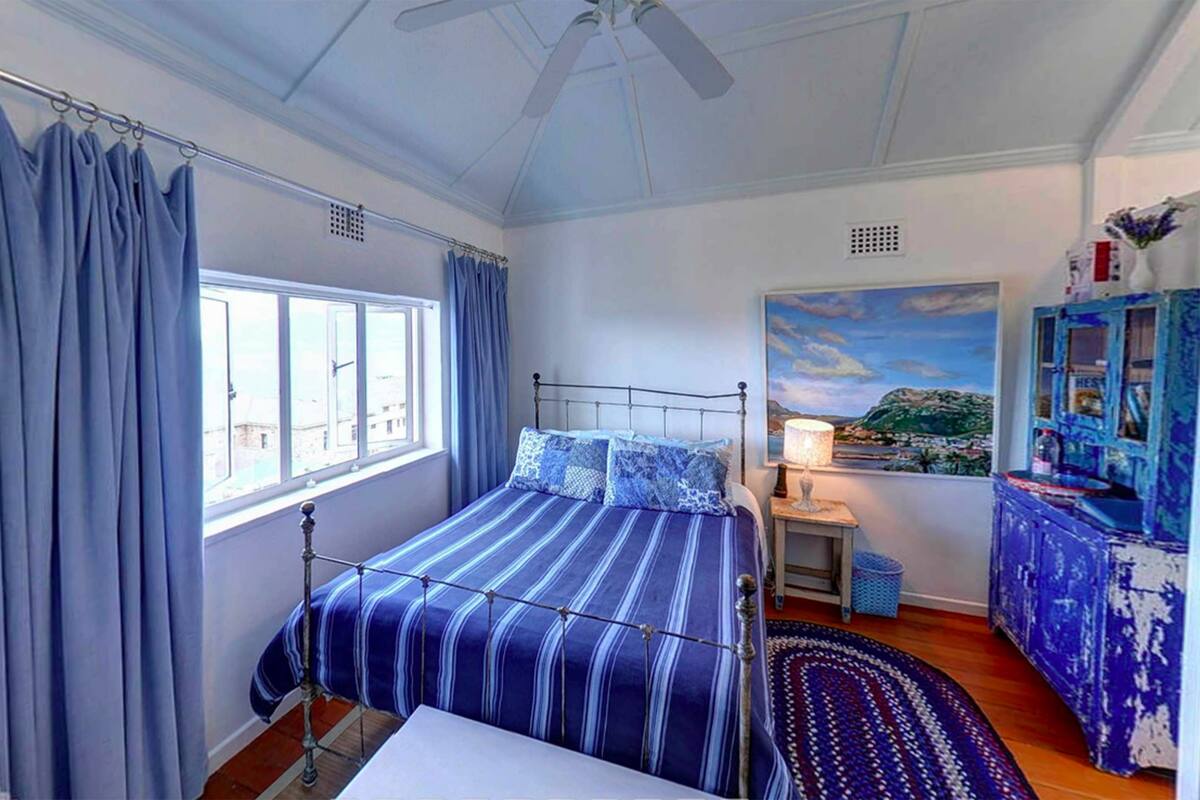 A bedroom with a bed, a nightstand, a lamp, a window with curtains, a ceiling fan, a painting, and a dresser.