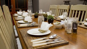 Een eettafel gedekt met witte borden, bestek en glazen, met een houten tafelkleed en potplanten.