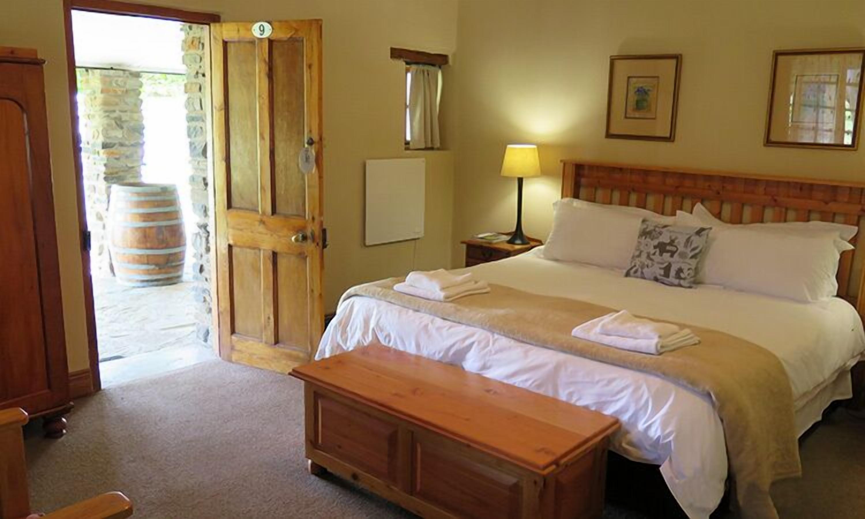 Habitación de hotel con cama de madera, una mesita de noche con lámpara, un banco de madera y un barril de piedra fuera de la puerta.