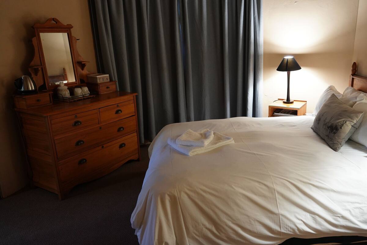 A hotel room with a bed, a wooden dresser, a nightstand with a lamp, and a painting on the wall.