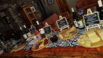 Una mesa con varios quesos, embutidos y botellas de vino, ubicada en una habitación rústica con paredes de piedra y muebles de madera.