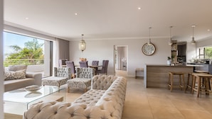 A modern living room with a large beige sofa, patterned armchairs, and a glass coffee table. There is a kitchen area with wooden stools and a large clock on the wall. - Sunrise Bay Self-Catering Accommodation (Plettenberg Bay)