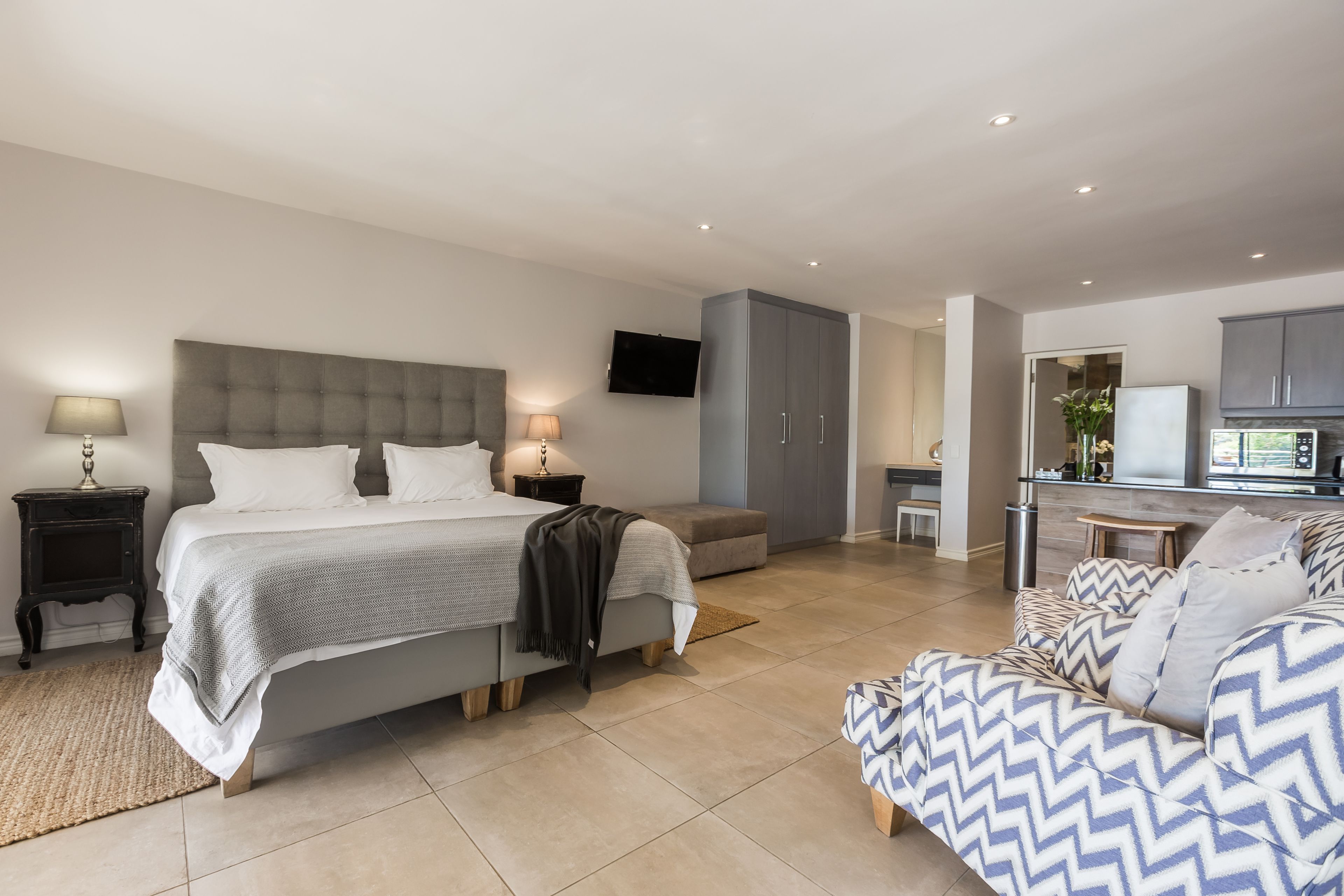 A modern bedroom with a large bed, bedside tables, a TV, and a kitchenette area.
